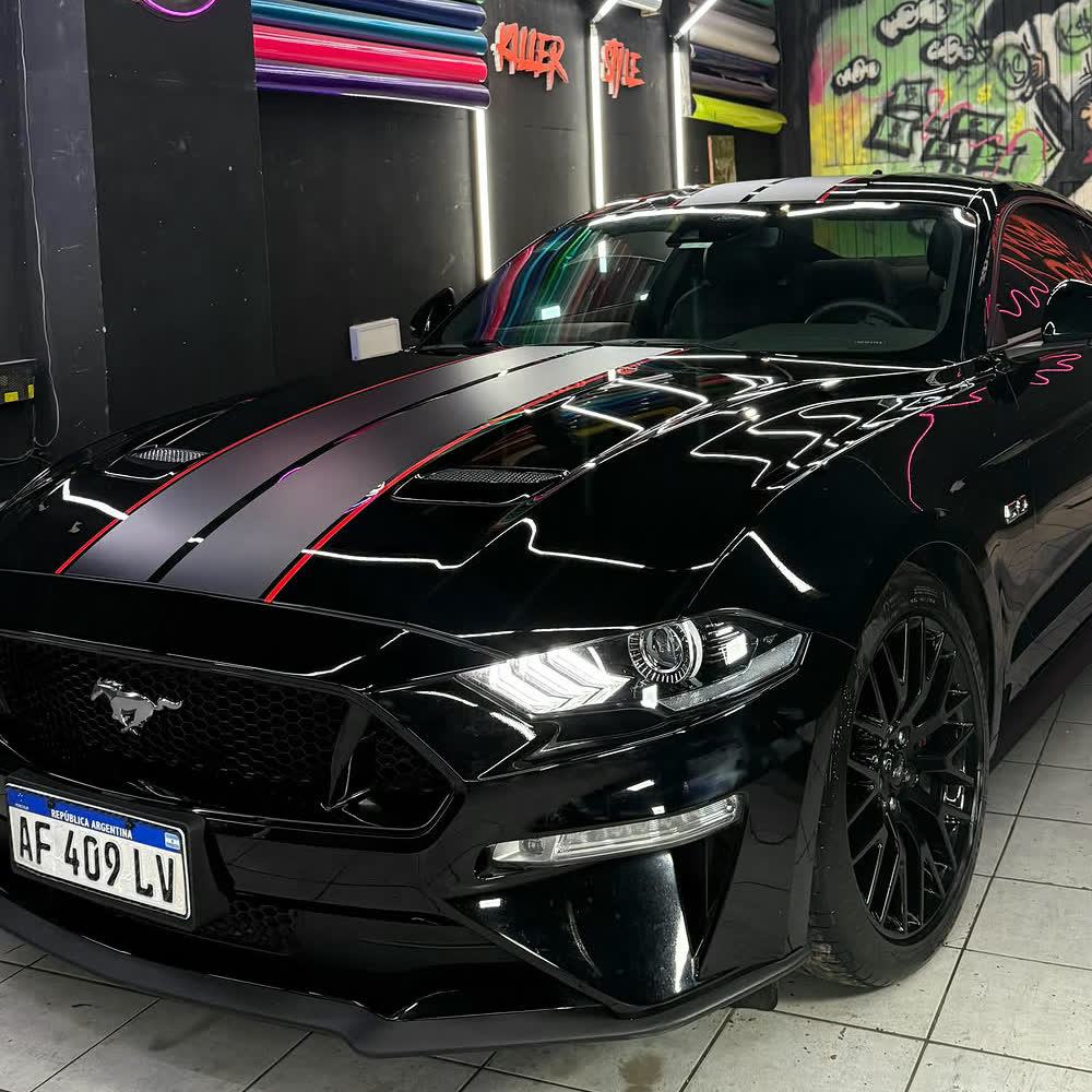 Un Ford Mustang negro con calcos en el taller de Killer Style, con el cartel de neón de la marca y los rollos de vinilo de colores en la pared